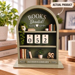 May include: An arched green wooden bookshelf display featuring the text "BOOKS Donated!" and "ALYSON'S LIBRARY." The unit includes three shelves decorated with miniature books, small potted plants, a decorative knot, and a flip-style counter showing 052.
