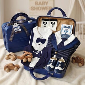 Puede incluir: Una maleta azul marino llena de ropa y accesorios para bebés. Incluye una camisa blanca con pajarita azul marino, un traje de raya diplomática azul marino, calcetines, zapatos y coches de juguete de madera. La maleta tiene el texto "LOVE IS ETERNAL" y "LITTLE MAN".