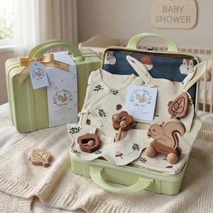 Op de afbeelding: Een babycadeauset in een lichtgroene koffer. De set bevat een crèmekleurige onesie, slab en hoed met een blad- en eikelpatroon. Houten speelgoed in de vorm van een blad, eikel en eekhoorn is ook inbegrepen. Een label luidt "Welkom op de wereld."