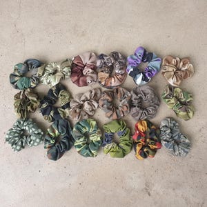 May include: A collection of colorful scrunchies in various patterns and colors, including green, blue, purple, and brown. The scrunchies are made of fabric and are arranged on a neutral surface. These hair accessories are perfect for adding a touch of style to any hairstyle.