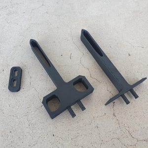 May include: Three black, geometric-shaped tools are arranged on a concrete surface. The tools include two T-shaped pieces and a small rectangular piece with two holes. The tools appear to be made of a matte black material.