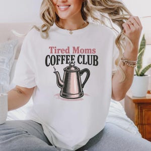 Può includere: T-shirt bianco con la scritta "Tired Moms COFFEE CLUB" in rosa e nero. Una grafica di caffettiera rosa e nera è centrata sotto il testo. Dalla caffettiera esce del vapore.