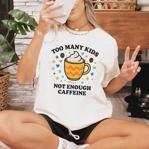 May include: Cream-colored t-shirt with the text "TOO MANY KIDS NOT ENOUGH CAFFEINE" and a graphic of a coffee cup with whipped cream. The design includes colorful stars, hearts, and coffee beans.