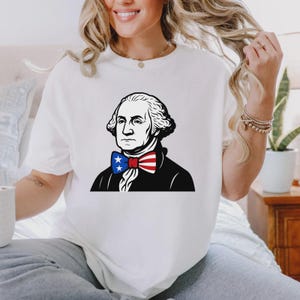 May include: White t-shirt with a graphic of George Washington wearing a bow tie in the style of the American flag. The illustration is in black and white, with the bow tie in red, white, and blue. The shirt is a casual style.