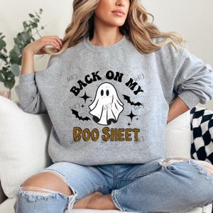 以下が含まれることがあります： 「BACK ON MY BOO SHEET」の文字が黒とオレンジでプリントされたライトグレーのスウェットシャツ。白いお化け、黒いコウモリ、星のデザインで、ハロウィーンに最適です。