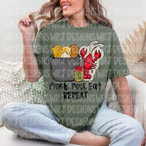 May include: Olive green t-shirt featuring a graphic of a pot with corn, lemon, and potatoes, a red crawfish, and a jar of seasoning. The text "Pinch, Peel, Eat, Repeat" is below the graphic. The shirt is a relaxed fit.