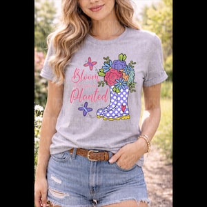 May include: A light gray t-shirt featuring a colorful graphic of a rain boot filled with flowers. The text "Bloom where you're Planted" is above the boot. The shirt is paired with denim shorts and a brown belt.