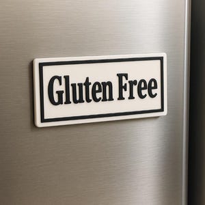 May include: A rectangular magnet with the words "Gluten Free" in bold black letters on a white background. The magnet has a black border and is affixed to a stainless steel surface.