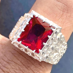 May include: A silver ring with a large, square-cut, deep red gemstone. The ring has a detailed, textured band with floral patterns. The gemstone is the focal point, catching the light and reflecting a vibrant color.