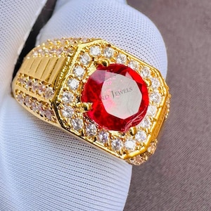 May include: A gold ring with a large, red gemstone at its centre, encircled by smaller, clear gemstones. The ring has a rectangular face and is set with multiple small stones. The text "ND JEWELS" is visible on the gemstone.