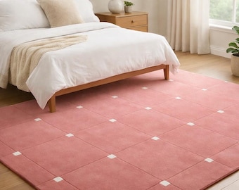 May include: A large, rectangular, dusty rose rug with a grid pattern of squares and white accent squares. The rug is placed in a bedroom setting, with a bed, nightstand, and lamp visible in the background.
