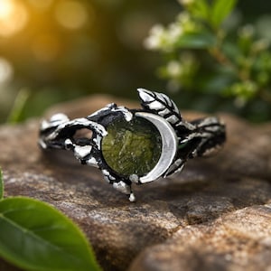 May include: A silver-toned ring featuring a textured band shaped like intertwined branches and leaves. The center of the ring holds a round, olive-green, textured stone partially framed by a smooth, polished silver crescent moon shape.