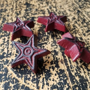 Puede incluir: Cuatro adornos de hierro fundido de color rojo en forma de estrella con un diseño de círculo negro en el centro. Los adornos están sobre una superficie de madera marrón oscura.