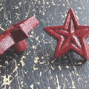 Può includere: Due pomelli a forma di stella di colore rosso scuro con un design a cinque punte. Un pomello è mostrato in prospettiva frontale, mentre l'altro è posizionato di lato, rivelando il profilo laterale e il punto di fissaggio cilindrico sul retro.