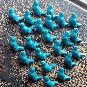 Puede incluir: Una colección de veinte pequeñas figuras con forma de pájaro de color verde azulado dispuestas en un patrón disperso. Cada figura presenta una superficie texturizada, un pico definido, un detalle de ala y una cola en un acabado uniforme.