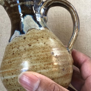 Könnte beinhalten: Handgefertigter Keramikkrug mit dunkelbraunem Oberteil und beigem Körper. Der Krug hat einen gebogenen Griff und einen Ausguss zum Ausgießen. Die Keramik hat eine gesprenkelte Textur und ein glänzendes Finish. Der Krug wird von einer Person gehalten.