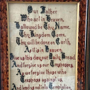 May include: Framed artwork featuring the Lord's Prayer, embroidered in black and red thread on a beige fabric. The wooden frame has a dark, patterned finish. The text is in an old English font.