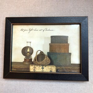 May include: Framed print featuring a still life with a kerosene lamp, a woven basket, and a stack of round boxes. The print includes the text "Let your light shine out of darkness..." in script. The frame is black.