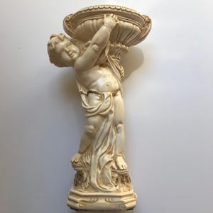 Op de afbeelding: Een crèmekleurig cherubijnbeeld dat een grote, sierlijke kom boven zijn hoofd houdt. De cherubijn is gehuld in vloeiende stof en staat op een decoratieve basis. Het beeld is gemaakt van een glad materiaal en heeft gedetailleerde kenmerken.