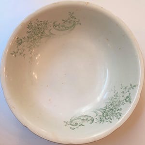 Może przedstawiać: Kremowa ceramiczna miska z falistym brzegiem. Misa zdobiona jest zielonym wzorem kwiatowym, z klastrami kwiatów i liści. Wzór powtarza się na powierzchni miski.