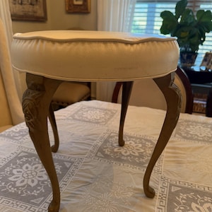Puede incluir: Un taburete de tocador antiguo con un asiento acolchado de color crema. El taburete tiene cuatro patas curvas con tallas ornamentadas y un acabado dorado. El taburete está colocado sobre un mantel de encaje blanco.