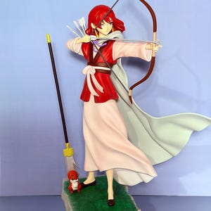 May include: A figurine of a character with red hair, dressed in a red and white outfit, holding a bow and arrow. The figure stands on a rock-like base with a small creature and a staff. The background is a solid light blue.