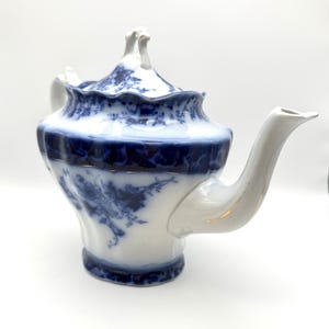 May include: A white porcelain teapot featuring a deep blue floral pattern. The teapot has a scalloped rim, a curved spout, and a lid with a decorative handle. The blue design is concentrated around the rim and base, with floral motifs.