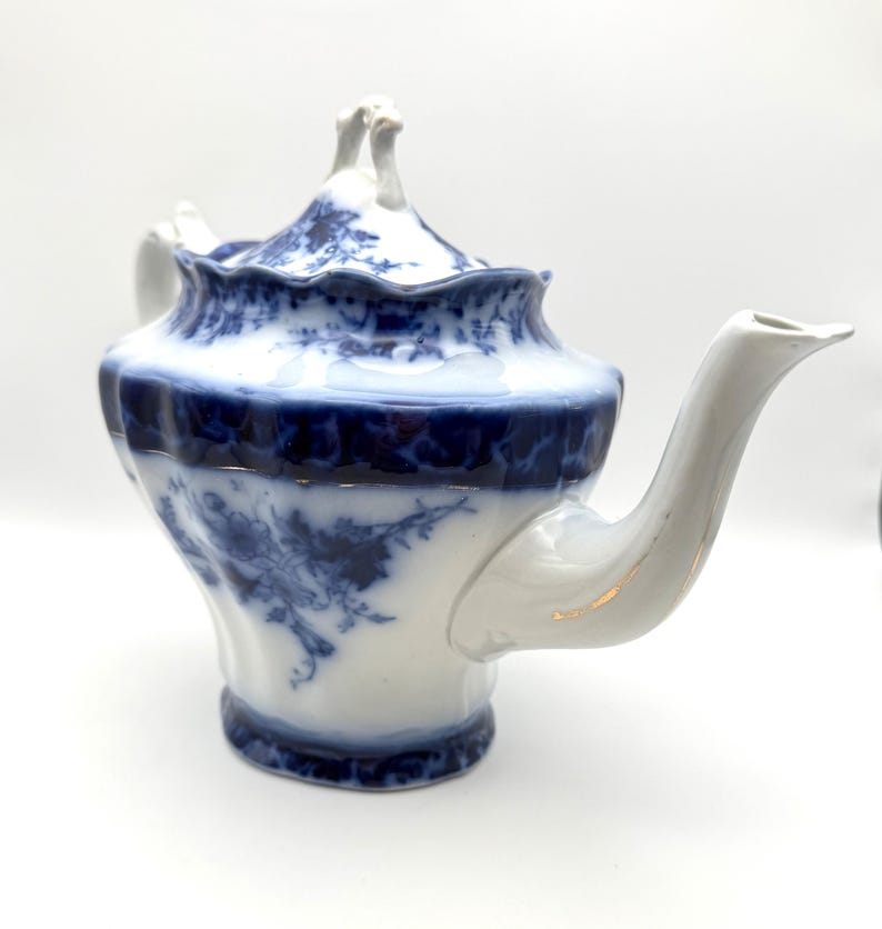 May include: A white porcelain teapot featuring a deep blue floral pattern. The teapot has a scalloped rim, a curved spout, and a lid with a decorative handle. The blue design is concentrated around the rim and base, with floral motifs.