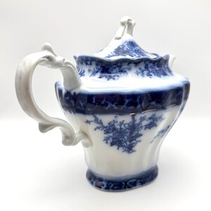 May include: A white porcelain teapot featuring a deep blue floral pattern and intricate scrollwork. The teapot has a scalloped rim, a curved handle with ornate detailing, and a matching lid with a decorative finial, all set against a plain background.
