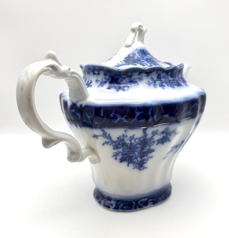 May include: A white porcelain teapot featuring a deep blue floral pattern and intricate scrollwork. The teapot has a scalloped rim, a curved handle with ornate detailing, and a matching lid with a decorative finial, all set against a plain background.