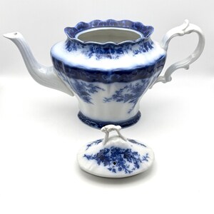 May include: A white porcelain teapot featuring a blue floral pattern and a scalloped rim. The teapot has a curved handle and a long spout. A matching lid with a small handle and a steam hole is placed in front of the teapot on a white surface.