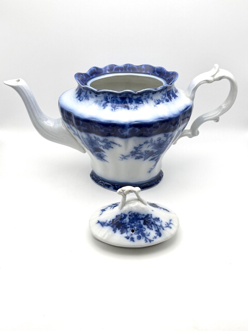 May include: A white porcelain teapot featuring a blue floral pattern and a scalloped rim. The teapot has a curved handle and a long spout. A matching lid with a small handle and a steam hole is placed in front of the teapot on a white surface.