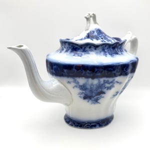 May include: A white porcelain teapot featuring a deep blue floral pattern and a scalloped rim. The teapot has a curved spout, a matching lid with a decorative finial, and a handle. The blue design includes intricate botanical motifs.