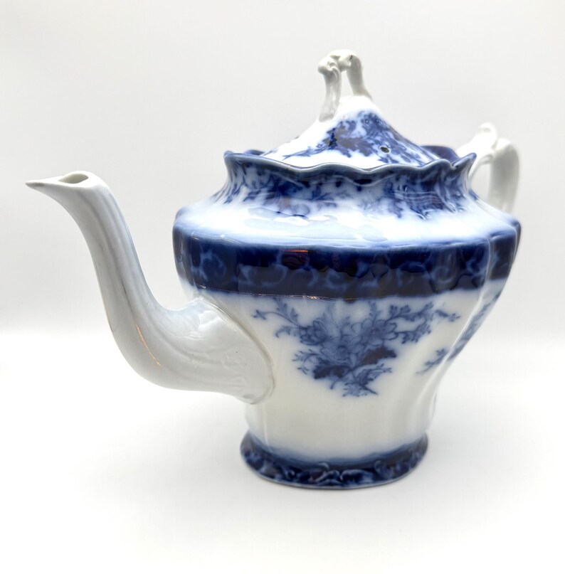 May include: A white porcelain teapot featuring a deep blue floral pattern and a scalloped rim. The teapot has a curved spout, a matching lid with a decorative finial, and a handle. The blue design includes intricate botanical motifs.