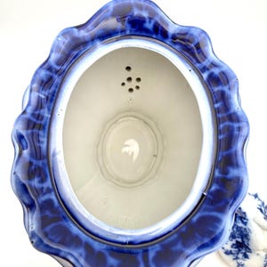 May include: A top-down view of a ceramic teapot with a scalloped rim featuring a deep blue and white mottled glaze. The interior is white with a built-in strainer containing five small holes near the spout. A separate matching lid sits nearby.