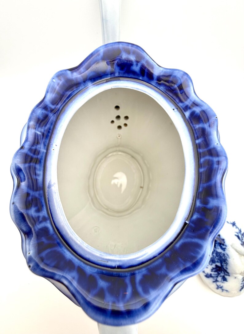 May include: A top-down view of a ceramic teapot with a scalloped rim featuring a deep blue and white mottled glaze. The interior is white with a built-in strainer containing five small holes near the spout. A separate matching lid sits nearby.