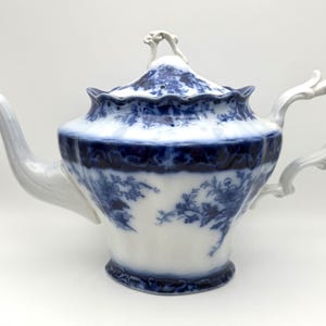 May include: A white porcelain teapot featuring a blue floral transferware pattern. The teapot has a scalloped rim, a curved handle, a tapered spout, and a matching lid with a decorative finial. The blue design covers the lid and upper body.