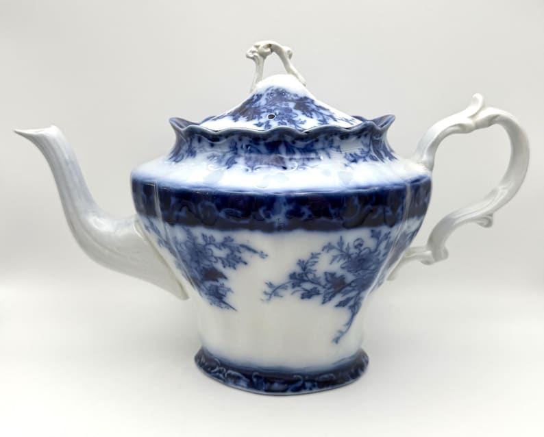 May include: A white porcelain teapot featuring a blue floral transferware pattern. The teapot has a scalloped rim, a curved handle, a tapered spout, and a matching lid with a decorative finial. The blue design covers the lid and upper body.