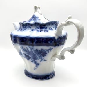 May include: A white porcelain teapot featuring a deep blue floral pattern. The teapot has a scalloped rim, a curved handle, and a matching lid with a decorative finial. The body displays intricate blue botanical illustrations against a white base.