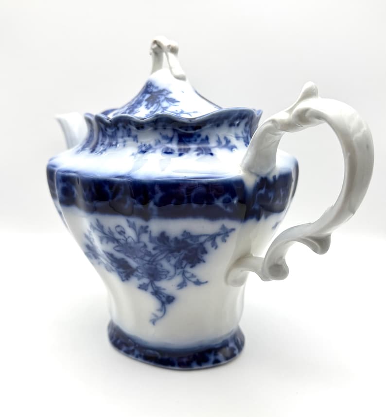 May include: A white porcelain teapot featuring a deep blue floral pattern. The teapot has a scalloped rim, a curved handle, and a matching lid with a decorative finial. The body displays intricate blue botanical illustrations against a white base.