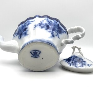 May include: A white porcelain teapot with a blue floral pattern and a matching lid. The base features a blue stamp reading TOURAINE STANLEY POTTERY ENGLAND. The teapot has a scalloped rim, a curved handle, and a spout, with the lid placed beside it.