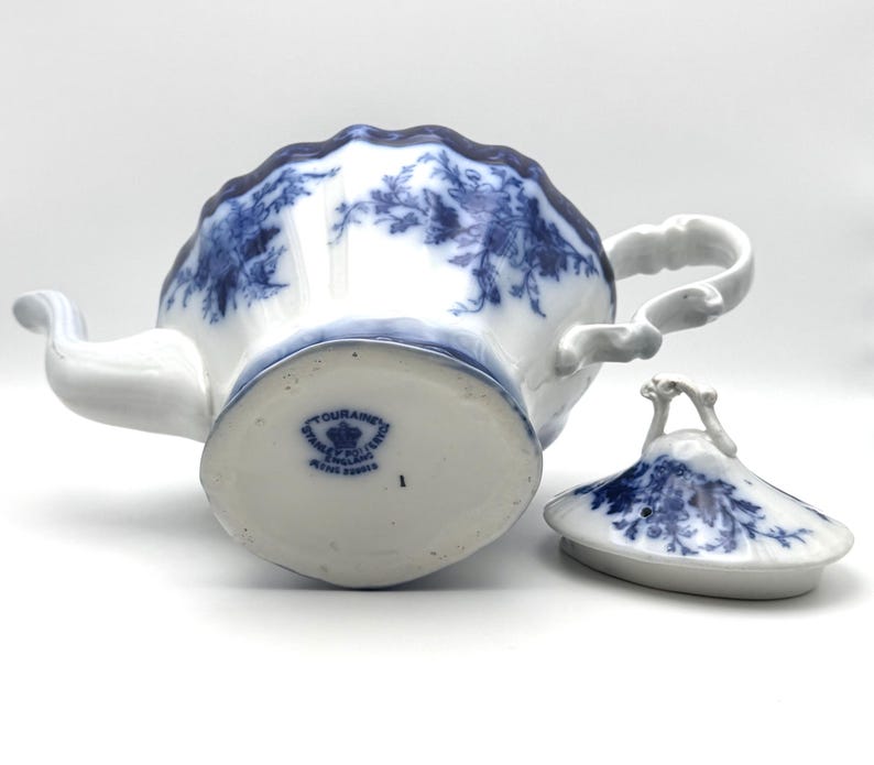 May include: A white porcelain teapot with a blue floral pattern and a matching lid. The base features a blue stamp reading TOURAINE STANLEY POTTERY ENGLAND. The teapot has a scalloped rim, a curved handle, and a spout, with the lid placed beside it.