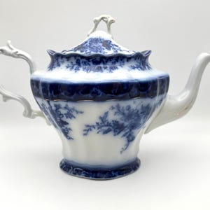 May include: A white porcelain teapot featuring a deep blue floral transferware pattern. The teapot has a scalloped rim, a curved handle, a tapered spout, and a matching lid with a decorative finial. The body displays intricate blue botanical motifs.