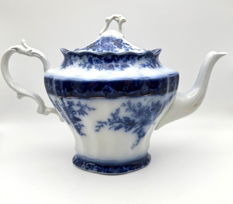 May include: A white porcelain teapot featuring a deep blue floral transferware pattern. The teapot has a scalloped rim, a curved handle, a tapered spout, and a matching lid with a decorative finial. The body displays intricate blue botanical motifs.