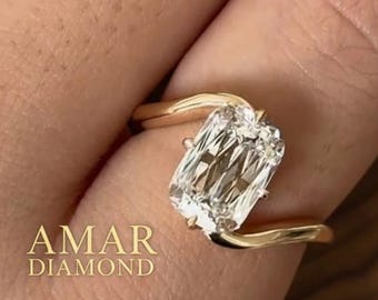 May include: A gold ring with a large, rectangular, clear diamond. The ring's band curves around the finger, and the diamond is set in a way that maximizes its brilliance. The text "AMAR DIAMOND" is visible.