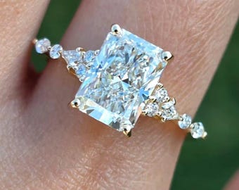May include: A radiant-cut diamond engagement ring with a gold band. The ring features a large rectangular center stone, flanked by smaller accent diamonds. Additional small diamonds are set along the band, creating a sparkling effect. The ring is shown on a finger.