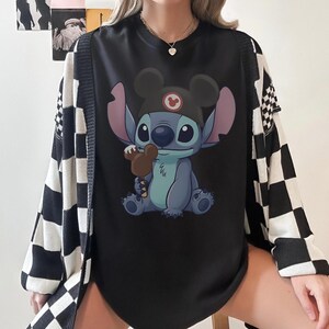 May include: Black graphic tee featuring Stitch from Lilo & Stitch wearing a Mickey Mouse hat and holding a brown ice cream bar. The tee is paired with a black and white checkered cardigan.