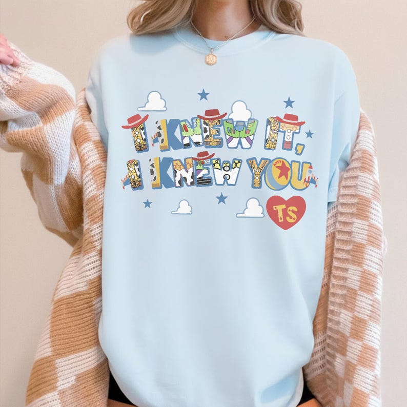 May include: A light blue crewneck sweatshirt featuring the text I KNEW IT, I KNEW YOU in a multi-colored font with patterns inspired by Toy Story characters, small red cowboy hats, blue stars, white clouds, and a red heart containing the letters TS.