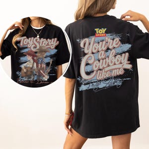 Puede incluir: Una camiseta negra con un gr&aacute;fico de Toy Story en la espalda con el texto "You're a cowboy like me" en letra grande. El dise&ntilde;o incluye un fondo de pincelada azul y el logotipo de Toy Story. Un recuadro circular muestra el gr&aacute;fico frontal.