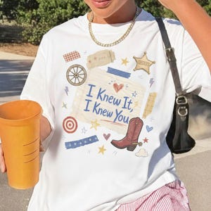 May include: A white t-shirt featuring a graphic print of a lined notebook paper with the text 'I Knew It, I Knew You' in blue script. The design includes a red cowboy boot, a sheriff star, a wagon wheel, and various small stars and hearts.